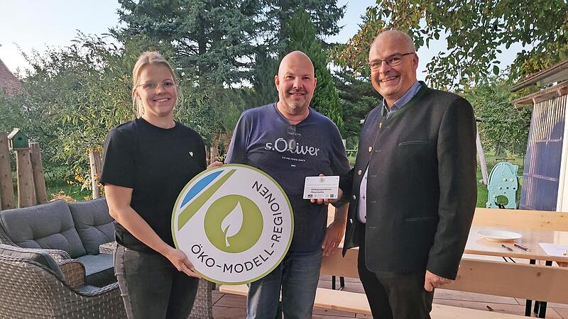 Die Öko-Modellregionmanagerin Laura Pircher und Gößweinsteins Bürgermeister Hanngörg Zimmermann (r.) übergeben die Förderplakette an Stefan Simmerlein.