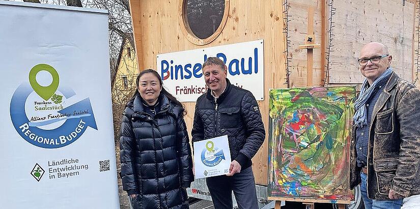 Allianzmanagerin Wenjun Zhang (v.l.), B&uuml;rgermeister Rainer Morper und Bernhard G&ouml;&szlig;mann-Schmitt vom Verein erLebenskunst freuen sich: Mit dem durch das Regionalbudget gef&ouml;rderten Anh&auml;nger bringt der Verein die Kunst in Schulen und Kinderg&auml;rten.