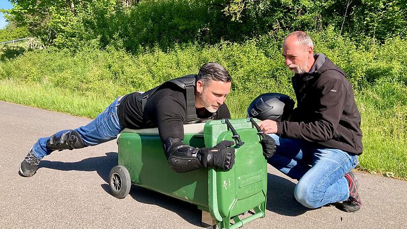 B&uuml;rgermeister Martin Finzel liegt auf einer gr&uuml;nen M&uuml;lltonne. Oliver V&ouml;lker weist ihn ein, bevor Finzel losf&auml;hrt.