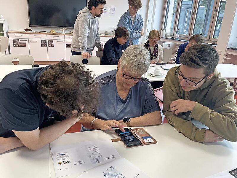 Zwei Sch&uuml;ler unterst&uuml;tzen eine Seniorin bei der Einstellung der Standortfreigabe auf ihrem Smartphone.