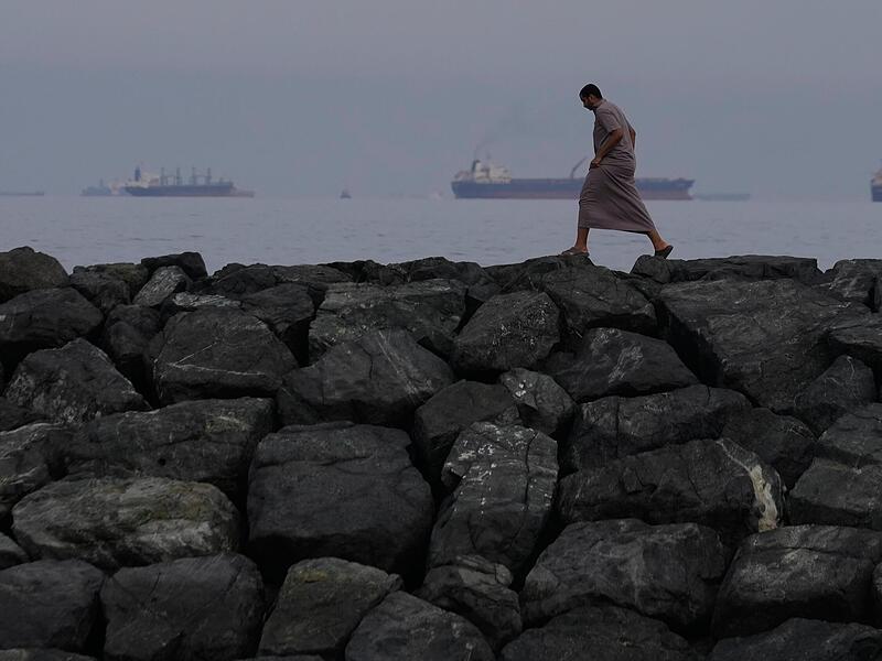 Ein Mann geht auf den Felsen am Ufer spazieren, w&auml;hrend &Ouml;ltanker und Frachtschiffe in der Stra&szlig;e von Hormus aufgereiht sind.