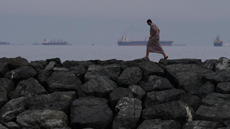 Ein Mann geht auf den Felsen am Ufer spazieren, w&auml;hrend &Ouml;ltanker und Frachtschiffe in der Stra&szlig;e von Hormus aufgereiht sind.