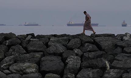 Ein Mann geht auf den Felsen am Ufer spazieren, w&auml;hrend &Ouml;ltanker und Frachtschiffe in der Stra&szlig;e von Hormus aufgereiht sind.