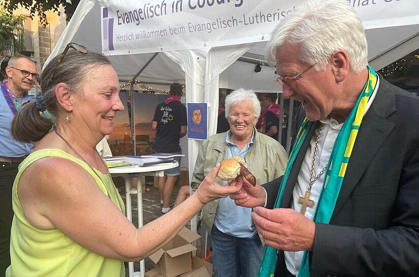 Landesbischof Heinrich Bedford-Strohm veröffentlichte dieses Foto auf seiner Facebook-Seite. Es zeigt, wie er von der Landessynodalen Petra Heeb eine Coburger Bratwurst überreicht bekommt.
