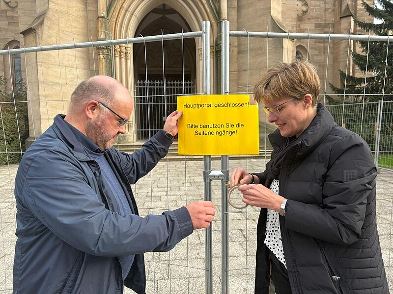 Pfarrer Gerd Greier und Gemeindereferentin Barbara Voll bringen das Hinweisschild am Bauzaun vor der Herz-Jesu-Kirche in Bad Kissingen an.
