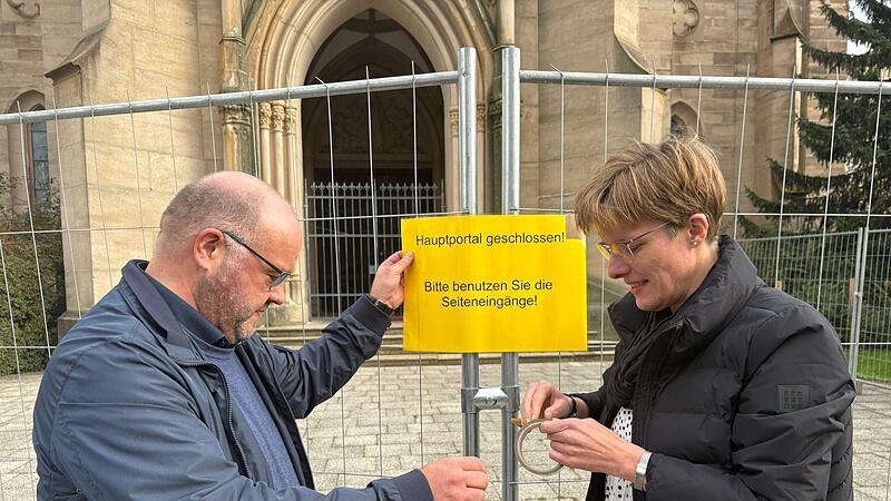 Pfarrer Gerd Greier und Gemeindereferentin Barbara Voll bringen das Hinweisschild am Bauzaun vor der Herz-Jesu-Kirche in Bad Kissingen an.