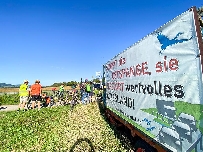 Die Fahrradgruppe hält neben einem ihrer Plakate bei Wiesenthau,  auf dem die Forderung steht: „Stoppt die Ostspange, sie zerstört wertvolles Ackerland“.