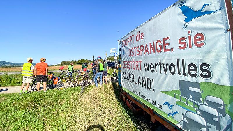 Die Fahrradgruppe hält neben einem ihrer Plakate bei Wiesenthau,  auf dem die Forderung steht: „Stoppt die Ostspange, sie zerstört wertvolles Ackerland“.