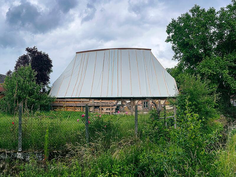 Nach Jahrzehnten des Verfalls wurde das historische Schwedenhaus in Poxdorf zun&auml;chst notgesichert. Die schweren Dachziegel wurden vollst&auml;ndig abgenommen, um das gesch&auml;digte Tragwerk zu entlasten. Eine gro&szlig;fl&auml;chige Schutzfolie bewahrt das Geb&auml;ude vor weiterem Wassereintritt.