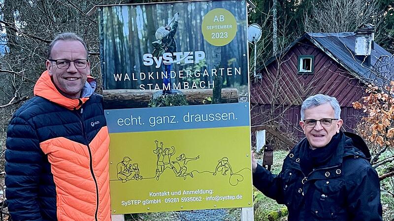 Michael Wilfert (links) vom Träger der Einrichtung und Bürgermeister Karl Philipp Ehrler mit der neuen Infotafel, die auf den entstehenden Waldkindergarten aufmerksam macht. Im Hintergrund die Waldhütte, die gerade vom Markt Stammbach saniert wird.