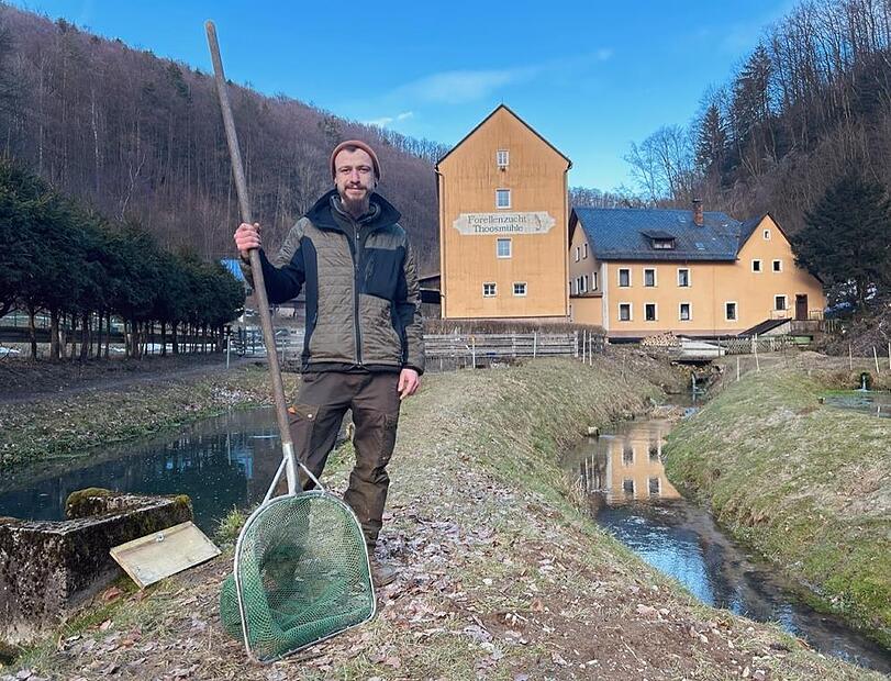 Daniel B&auml;rthlein steht mit einem Kescher an den Fischbecken und dem Bach vor der Thoosm&uuml;hle.