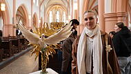 „Der trügerische Glanz“ nennt Annika Demmert ihr Werk. Eine weiße Taube, das universelle Friedenssymbol, steigt aus einem goldenen Kranz voller Flammen hervor: Doch die Kritik erschließt sich den Betrachtern von der anderen Seite. Denn in der Taube steckt ein Dolch in Kreuzform.