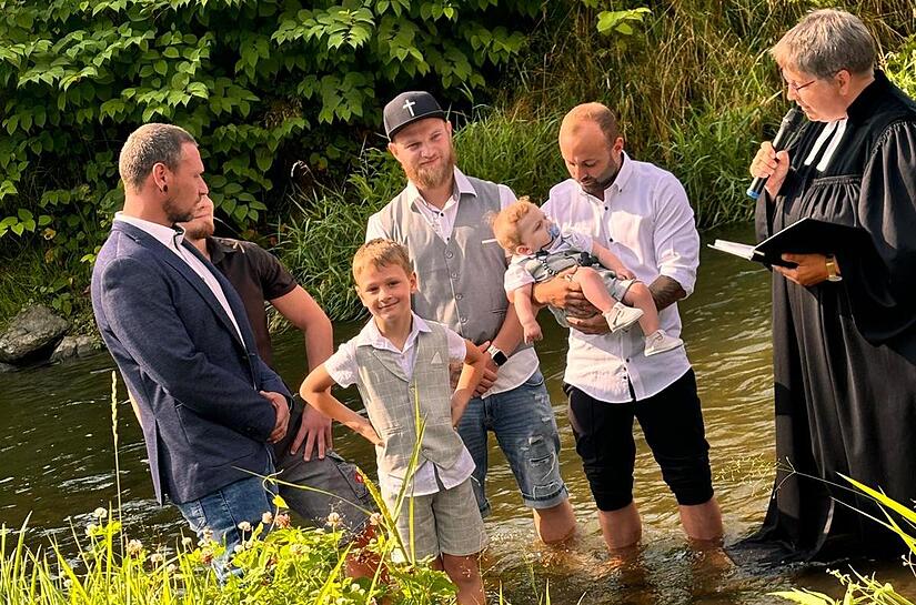 Pfarrerin Godila Baumann (rechts) stand mit ihrem schwarzen Talar ebenso im Wasser wie die Tauf-Familie mit den beiden T&auml;uflingen Tyler und Jayden (mit Schnuller).