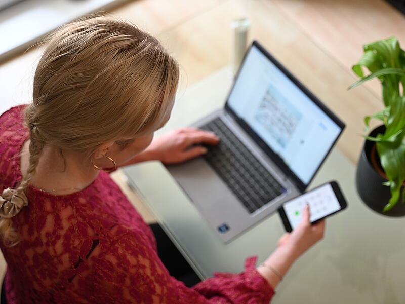 Frau sitzt vor Laptop und schaut auf Smartphone