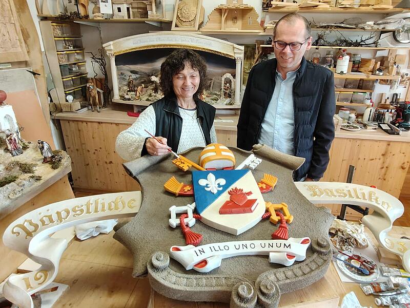 Lisa Schinner (hier mit Pfarrer Pater Florian) stellt mit letzten Pinselstrichen das neue Wappenschild von Papst Leo XIV. für die Basilika Marienweiher fertig.
