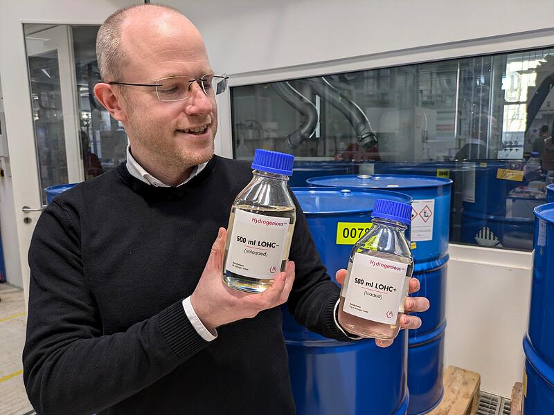 Frank Erik Walter zeigt die zwei Varianten der Tr&auml;gerfl&uuml;ssigkeit f&uuml;r Wasserstoff. Links die Variante ohne Wasserstoff, die leicht gelblich erscheint. Rechts die geladene Variante mit Wasserstoff, die komplett farblos aussieht.