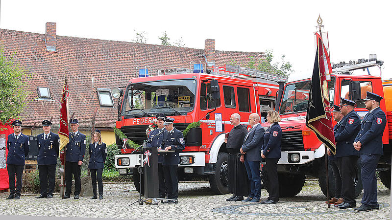 Bei der Fahrzeugweihe: am Rednerpult in der Mitte Kreisbrandmeister Alexander Frey (links) und der stellvertretende Kommandant der Münnerstädter Wehr, Guido Denner (rechts). Rechts daneben Pater Markus Reis, Bürgermeister Michael Kastl und die Kreisfrauenbeauftragte Birgit Below. Links und rechts zwei Fahnen-Abordnungen.
