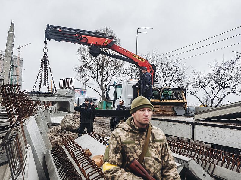 Arbeiter laden Betonplatten, w&auml;hrend ein bewaffneter ukrainischer Mann Wache steht. Russland hat seinen Angriff auf die zweitgr&ouml;&szlig;te Stadt der Ukraine fortgesetzt, obwohl beide Seiten erkl&auml;rt haben, dass sie bereit sind, die Gespr&auml;che wieder aufzunehmen.