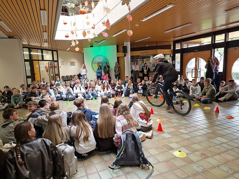 Landrat Mario G&ouml;tz f&auml;hrt unter dem Applaus der Sch&uuml;lerinnen und Sch&uuml;ler mit dem Fahrrad durch die Aula zur offiziellen Ehrung.