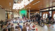 Landrat Mario G&ouml;tz f&auml;hrt unter dem Applaus der Sch&uuml;lerinnen und Sch&uuml;ler mit dem Fahrrad durch die Aula zur offiziellen Ehrung.