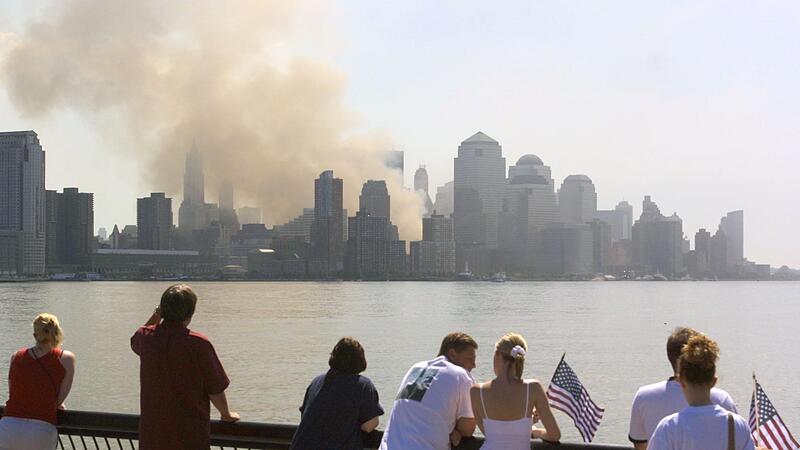 Passanten mit US-Fahnen betrachten am 12. September 2001 von New Jersey aus die in Rauch und Staub geh&uuml;llte Skyline von Manhattan. Dort wo am Vortag noch die beiden T&uuml;rme des World Trade Centers gestanden hatten, ist jetzt nur noch eine Qualmwolke zu sehen.