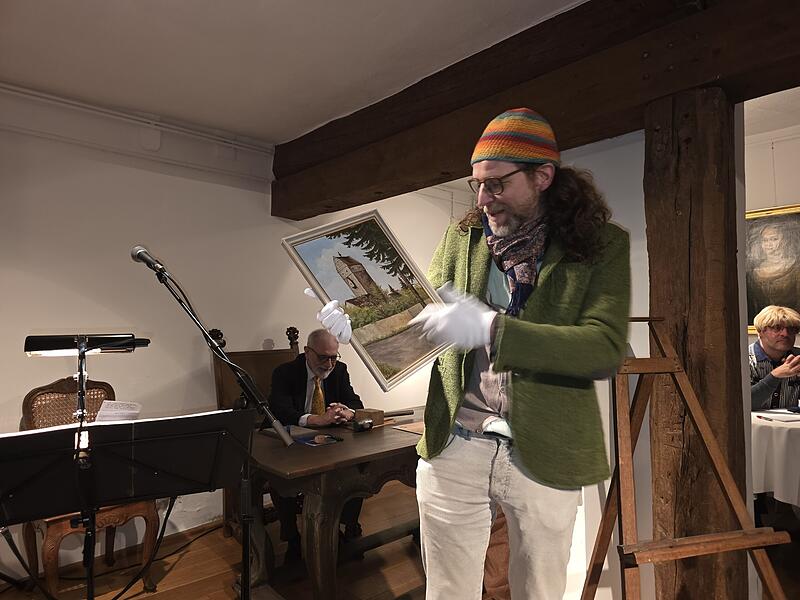 Dr. Nicolas Zenzen alias Prof. Dr. Zensorius mit einem &Ouml;lgem&auml;lde, das das Obere Tor zeigt. Im Hintergrund Auktionator Leo Pfennig (links) und Oliver Schikora am Bietertelefon..