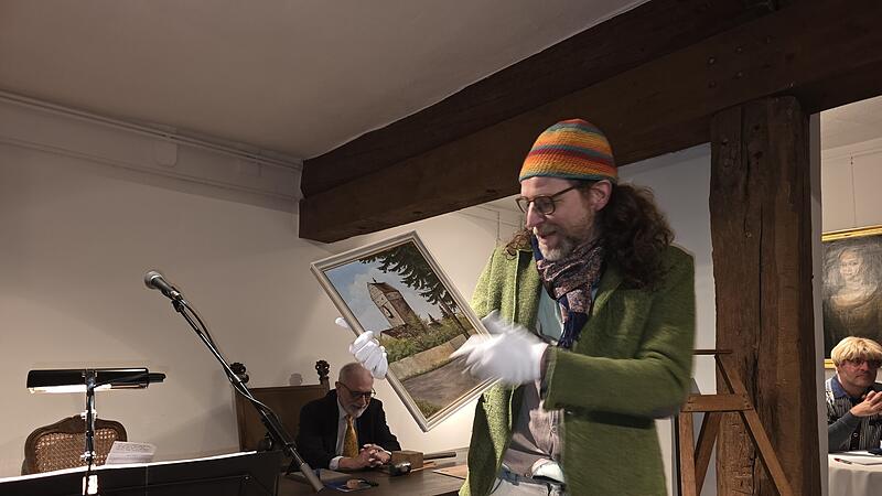 Dr. Nicolas Zenzen alias Prof. Dr. Zensorius mit einem Ölgemälde, das das Obere Tor zeigt. Im Hintergrund Auktionator Leo Pfennig (links) und Oliver Schikora am Bietertelefon..