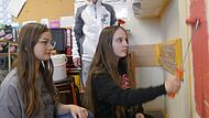 Neco Gülmen vom Malerfachbetrieb Scholten zeigt Jule und Marlene, wie Farbe an die Wand der Mittelschule Liebfrauenhaus kommt.