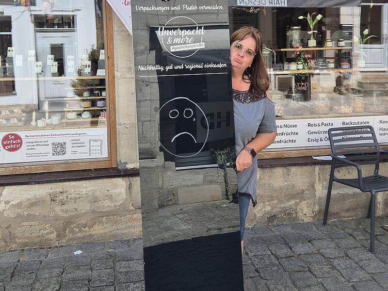 Alexandra Lowig zeigt vor ihrem Unverpacktladen das Schild, das sie aus Kostengr&uuml;nden nicht mehr aufstellen kann.