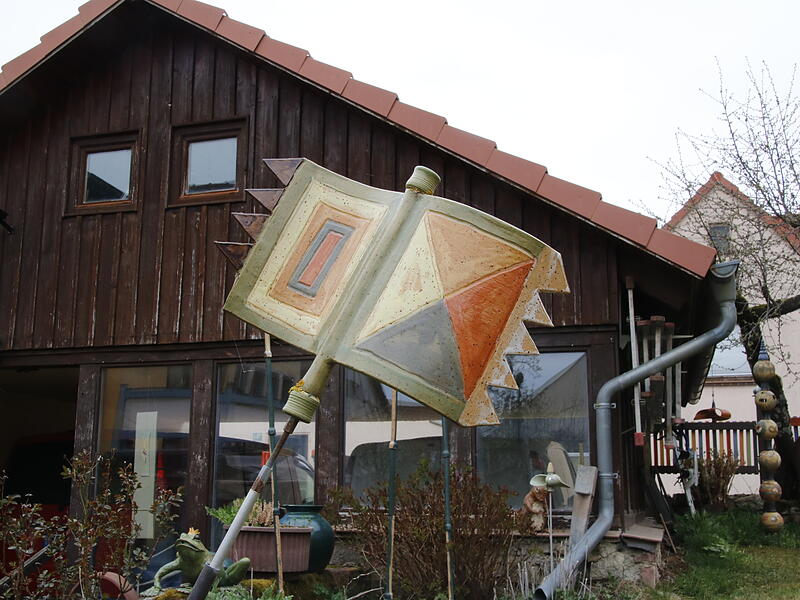 Eine Keramikflagge aus Erdt&ouml;nen steht vor einer kleinen Scheune im Garten.