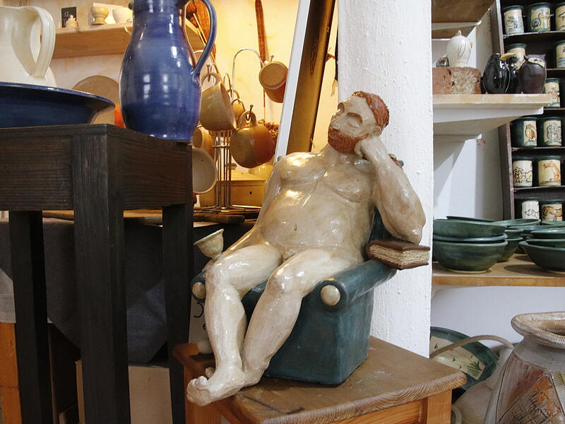 Keramikfigur, die einen nackten, b&auml;rtigen Mann darstellt, der entspannt auf einem Sessel sitzt.