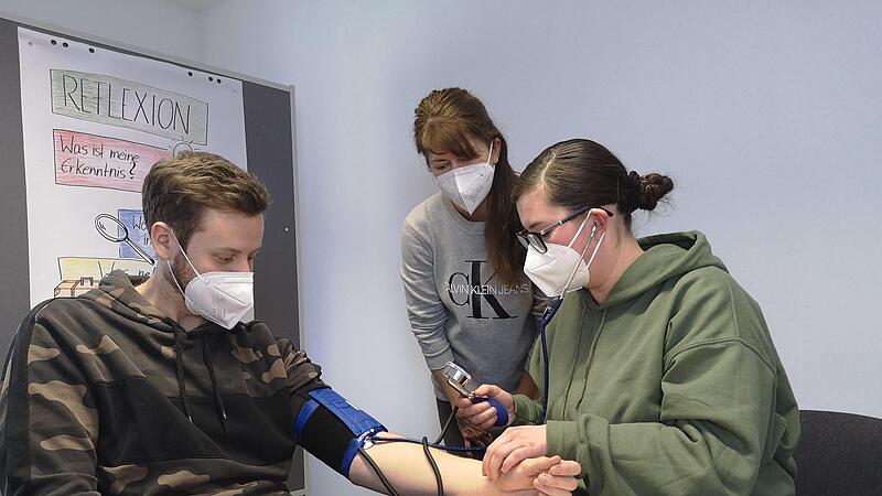 Anna Maria Trager (rechts) übt das Blutdruckmessen an Valentin Rau, Auszubildender im Diakonie-Seniorenzentrum Hephata, im Skills Lab der Diakoniestation Drei-Franken-Eck unter den wachsamen Augen von Praxisanleiterin Michaela Wagner.