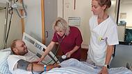 Schwester Eva (rechts) beobachtet, wie Sofia einem Patienten im Krankenhaus in H&ouml;chstadt den Blutdruck misst.