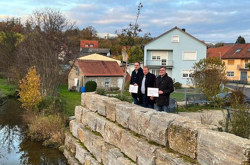 Am Rande der Aurach in Kirchaich, die renaturiert werden soll, übergab der Leiter des Amtes für ländliche Entwicklung Unterfranken, Jürgen Eisentraut (Mitte) den Förderbescheid an Bürgermeister Thomas Sechser (rechts) sowie an Rauhenebrachs Bürger...