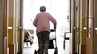 Eine Frau geht mit einem Rollator in einem Pflegeheim den Gang entlang.