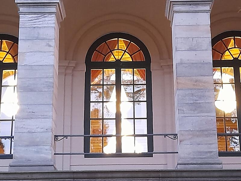 Während Musiker des Bayerischen Kammerorchesters bei Beleuchtung mittels eindrucksvoller Lüstern im Kursaalgebäude fürs nächste Konzert proben, genießen Passanten neben dem historischen Gebäude einen schönen Sonnenuntergang.