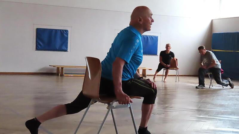 &Uuml;bung mit Stuhl: Oberk&ouml;rper vorschieben, Ferse nach hinten dr&uuml;cken, kurz halten, l&ouml;sen.